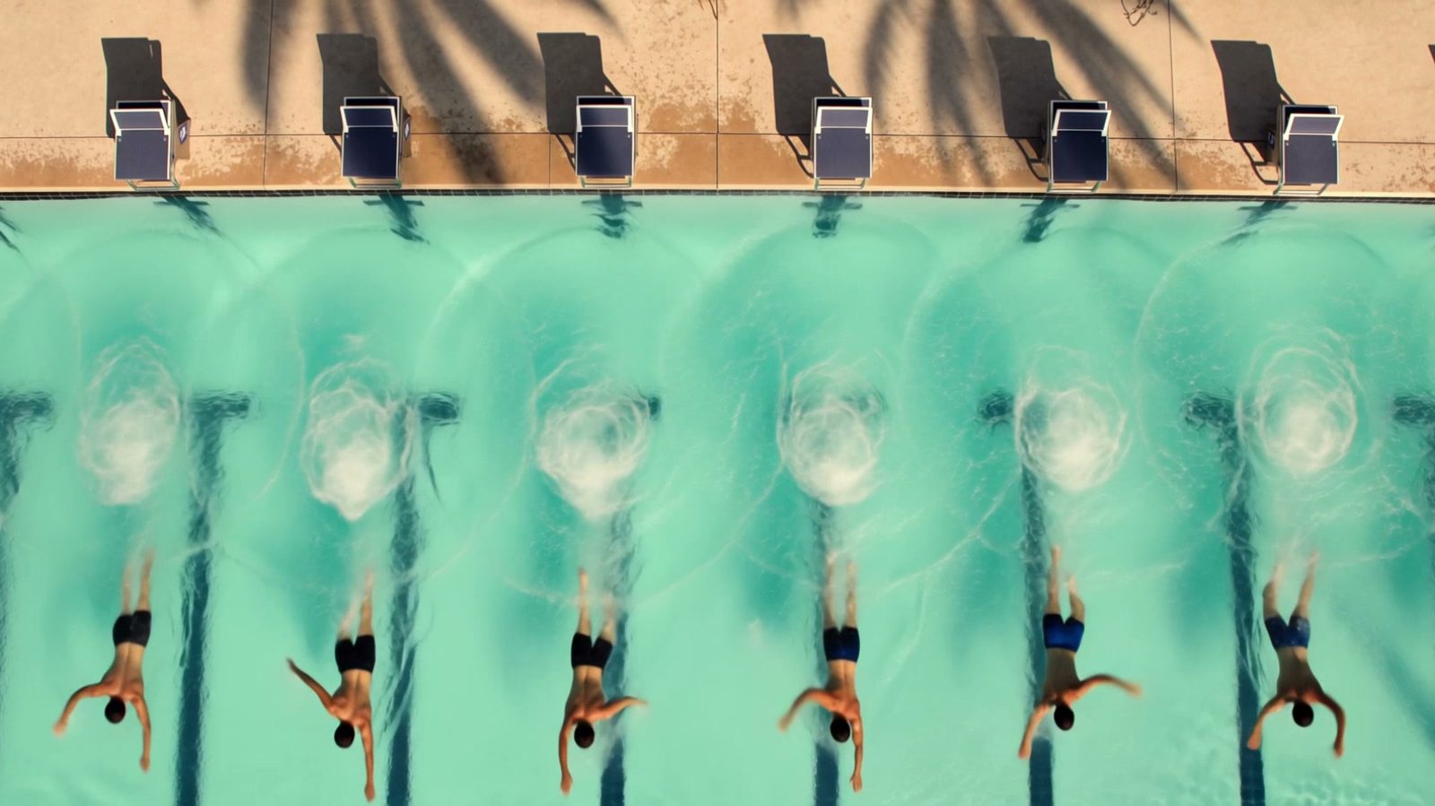 Kling 4.0 still: overhead view of an outdoor pool as eight swimmers glide underwater in a line after diving from the blocks, palm shadows on the deck.