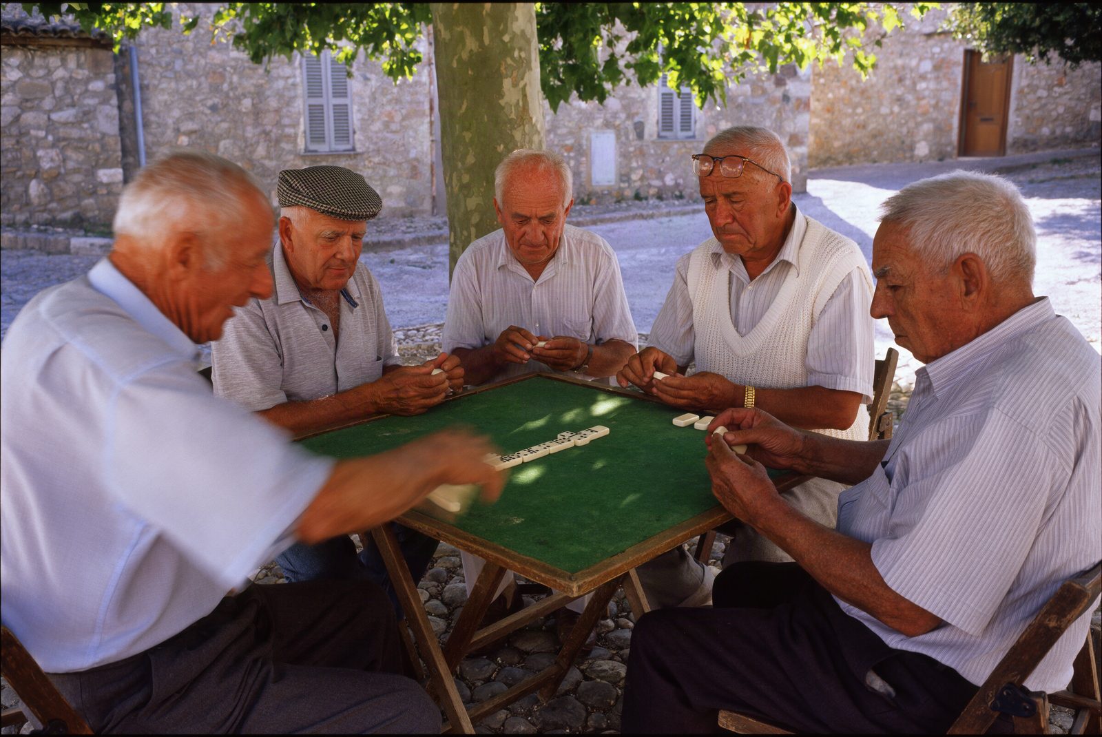 Krea 3: five elderly men playing dominoes on green felt under a plane tree, the man on the left slamming down a motion-blurred tile.