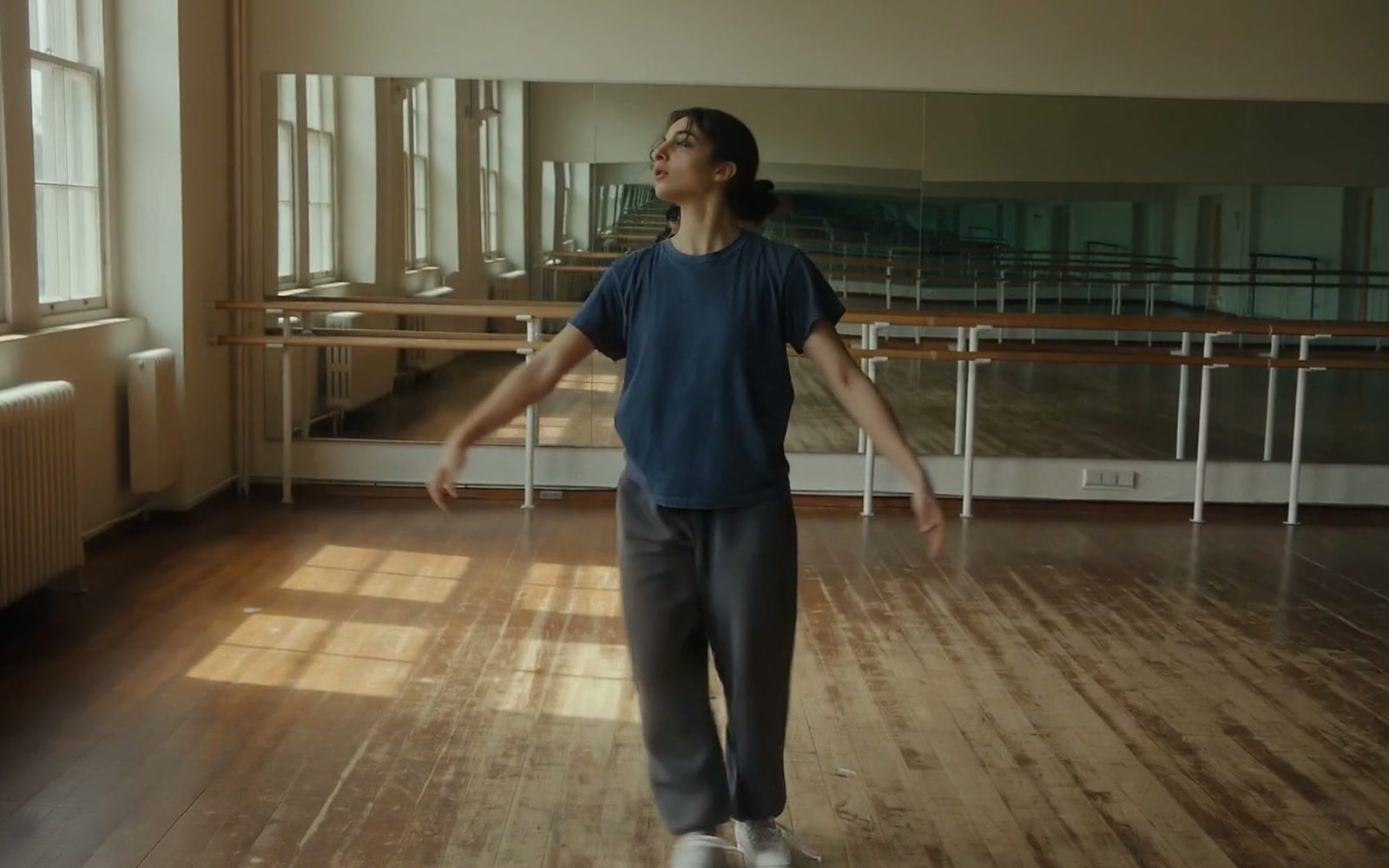 FLUX.3 Video still: a young dancer in a navy T-shirt and grey sweatpants turns with her arms open in a sunlit wooden rehearsal studio.
