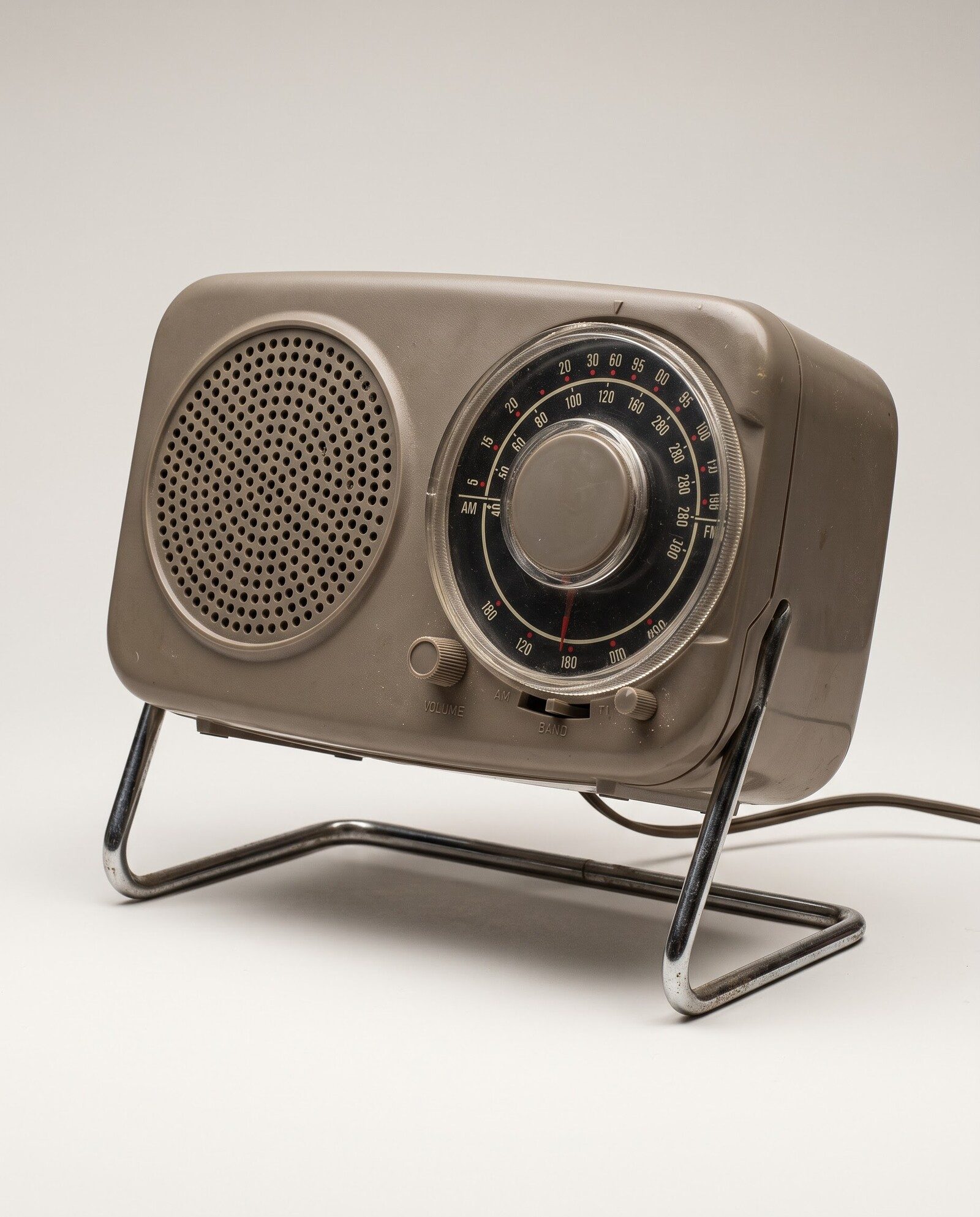 Reference photo for the edit test: a warm grey 1960s tabletop radio with a black tuning dial on a chrome tube stand, on a plain studio backdrop.