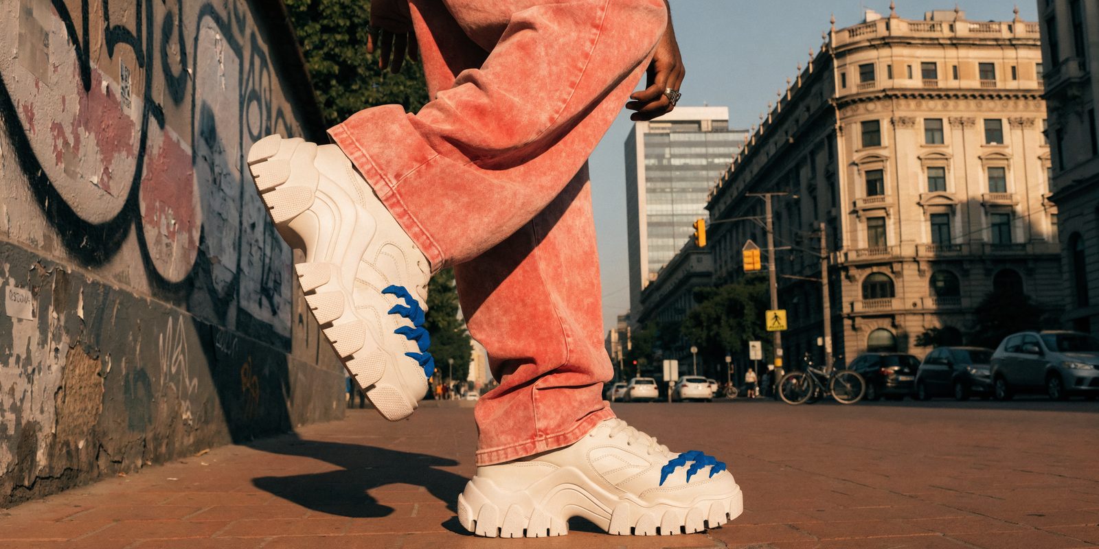 Lifestyle footwear concept showing chunky white sneakers and coral trousers beside a graffiti-covered city wall.