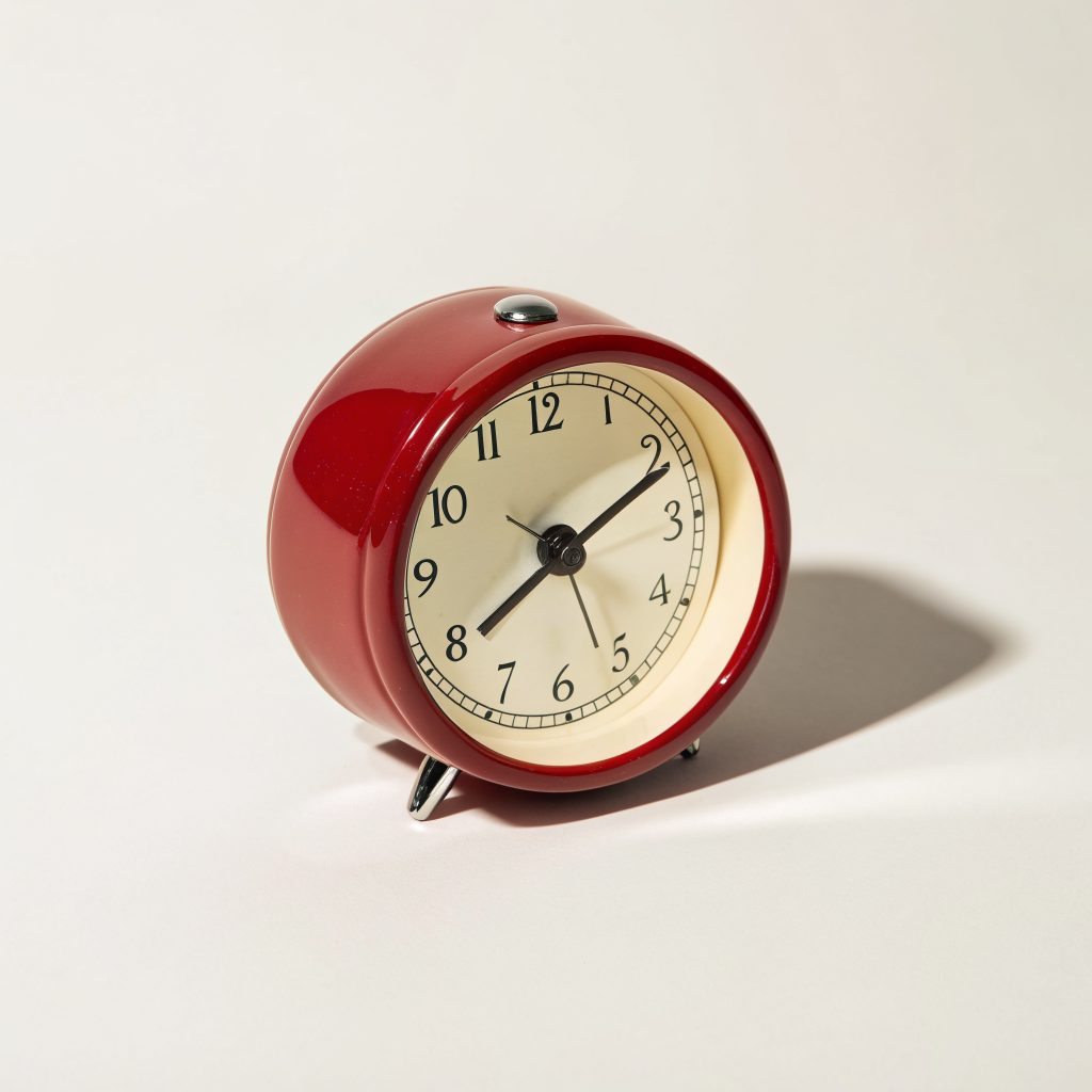 Red enamel alarm clock centered against an ivory background with a soft cast shadow.