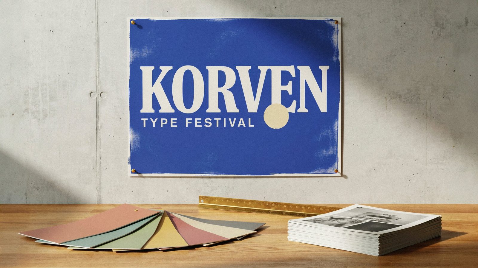 Cobalt screen-printed poster reading KORVEN and TYPE FESTIVAL pinned to a concrete wall above an oak desk with paper swatches, a brass ruler and printed proofs
