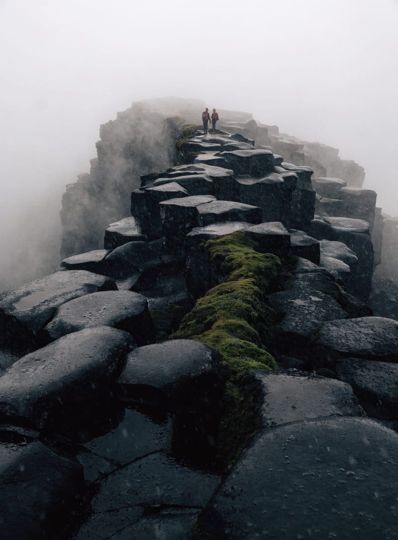 Fictional reportage scene of two distant hikers on a misty volcanic ridge.