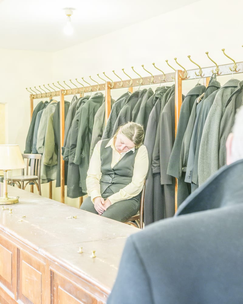 Cloakroom attendant in a waistcoat asleep on a stool among rows of olive wool coats, seen over a waiting patron's shoulder