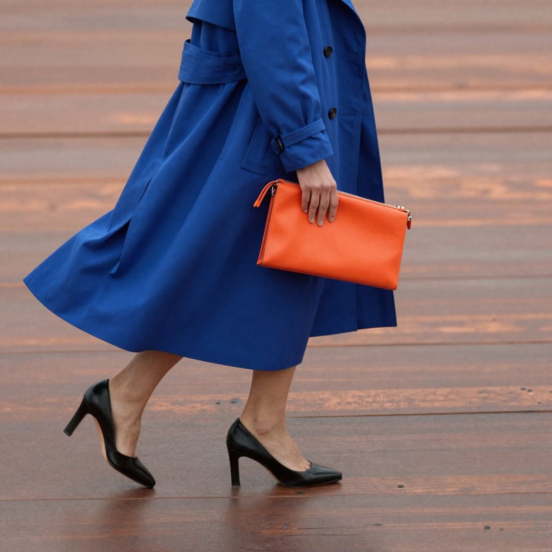 Fashion concept still of a person in a flowing blue coat carrying an orange clutch while walking across wet sand.