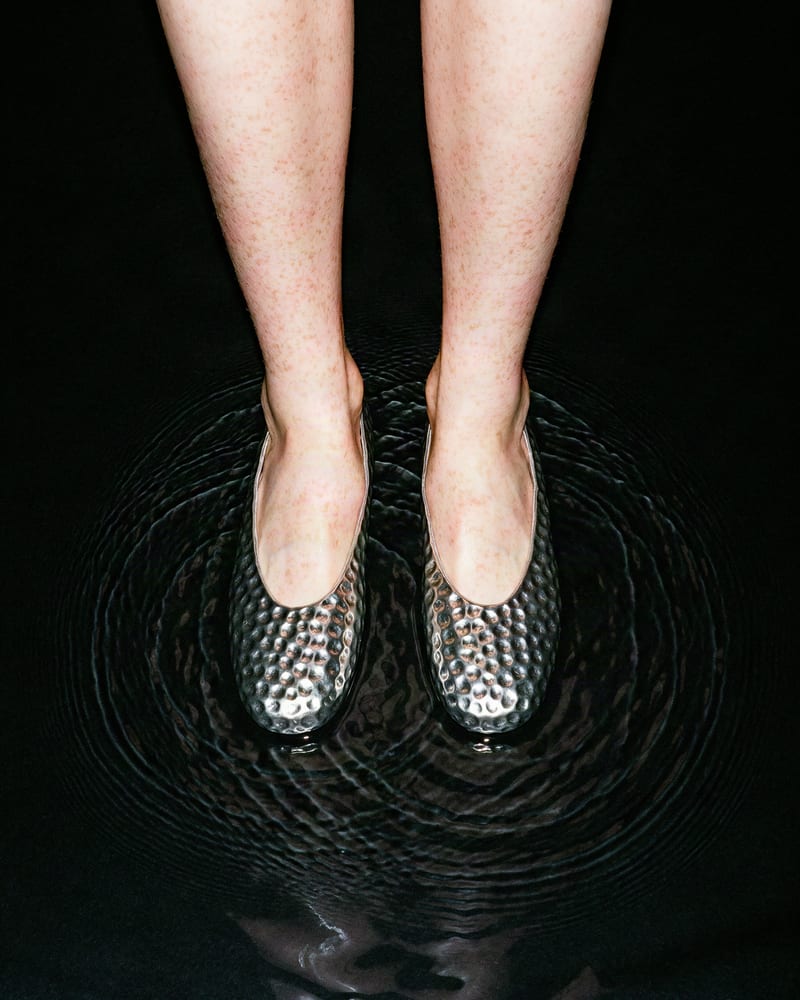 Overhead view of pale freckled shins in hammered pewter ballet flats standing in black water, ripples circling the feet