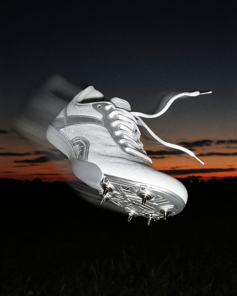 Single white-and-silver track spike frozen mid-air in camera flash, laces whipping and a motion-blur trail behind it, against an orange dusk band over a dark field