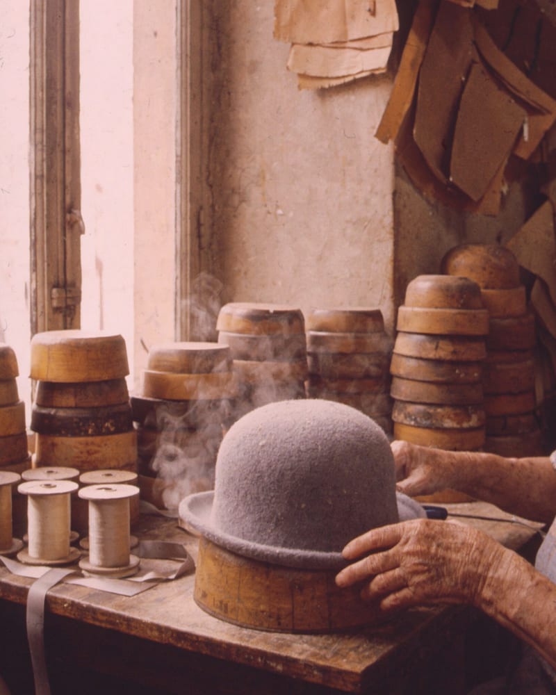Faded 1970s photo of an elderly milliner's hands steaming a grey felt hat on a wooden block by a window