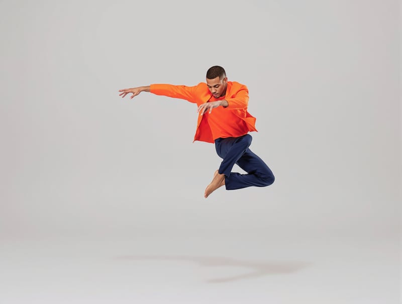 Fashion editorial of a dancer in an orange jacket and navy trousers jumping in a pale studio.