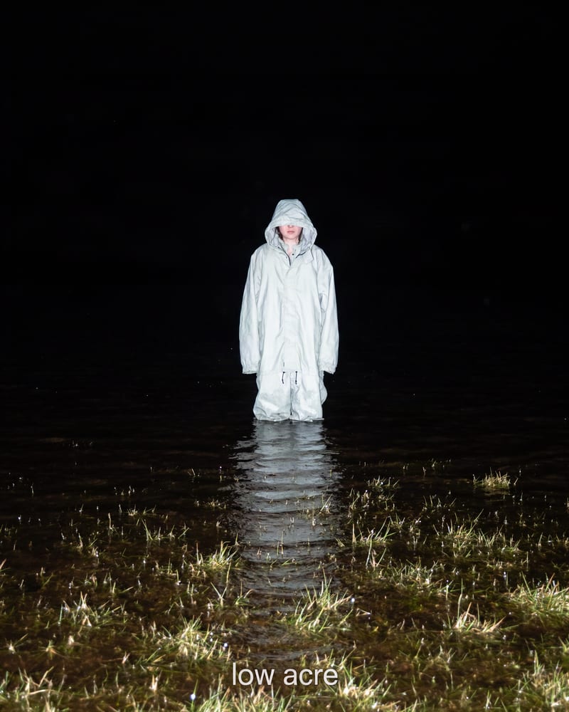 Album cover: flash-lit figure in a pale hooded anorak standing knee-deep in a flooded field at night, with the words low acre