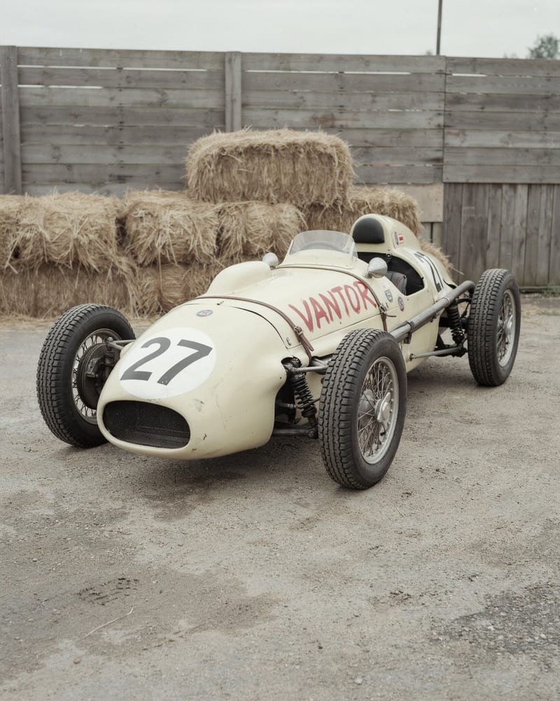 Faded film-style photo of a cream vintage open-wheel race car with red brush lettering VANTOR and number 27 in a white roundel, hay bales behind
