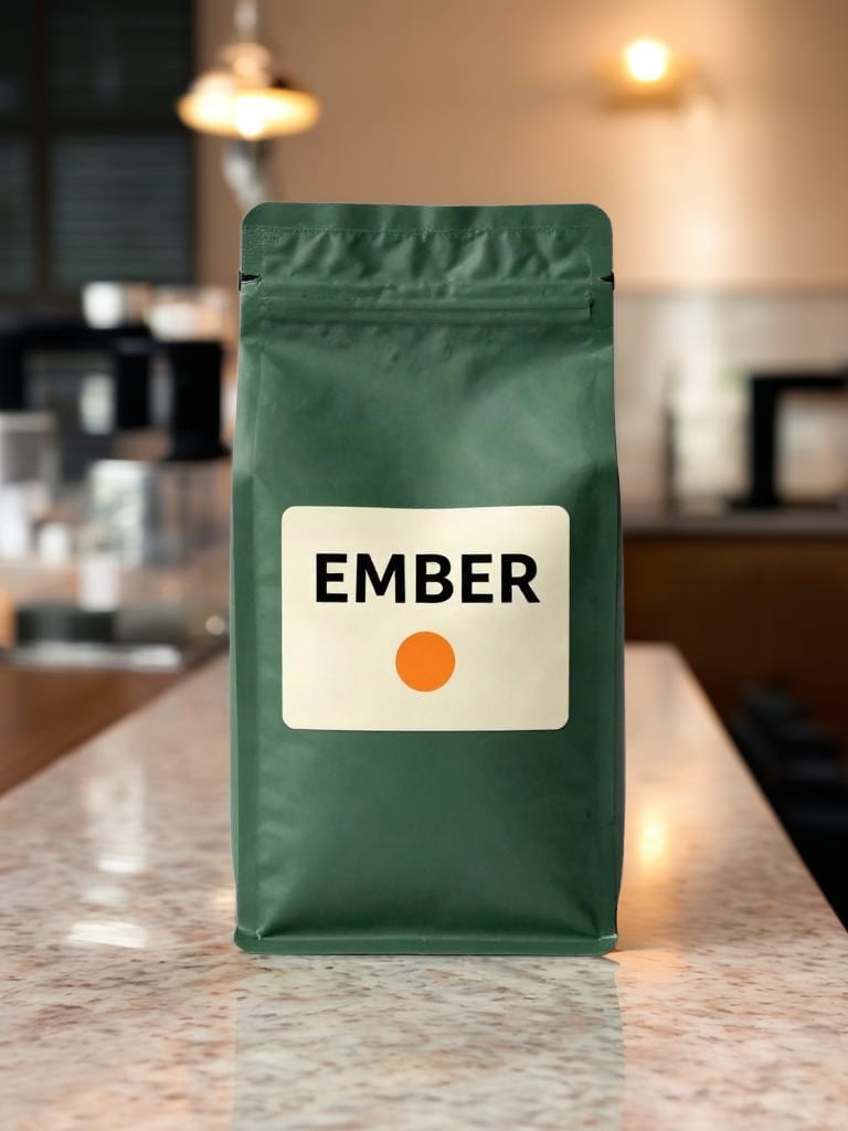 Green EMBER coffee bag retaining its cream label and orange dot on a cafe counter.