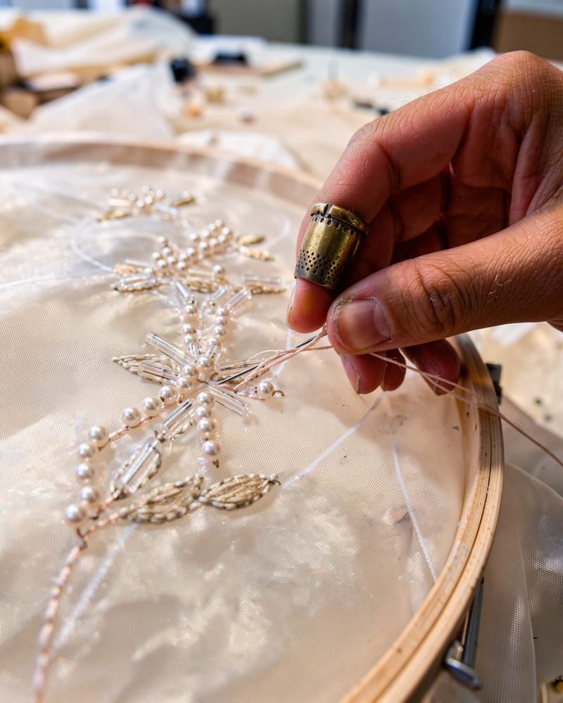 Macro of a couturier's thimbled hand stitching seed pearls and glass bugle beads onto ivory silk organza in an embroidery hoop