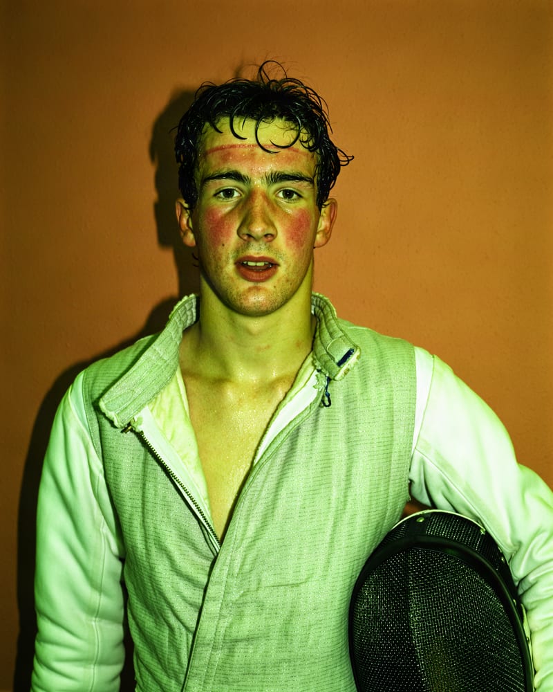 Sweaty young fencer with a red mask line on his forehead, jacket unzipped, mesh mask under his arm, in cross-processed strobe light