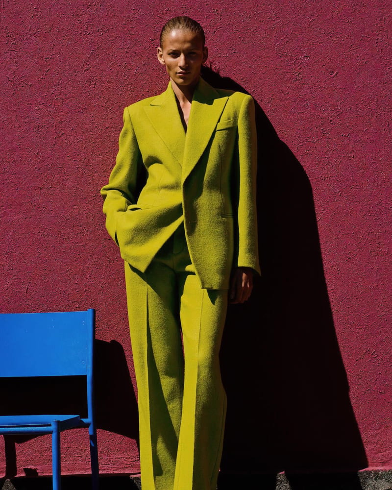 Model in an acid-green wool suit against a magenta stucco wall in hard sun, beside a cobalt-blue chair