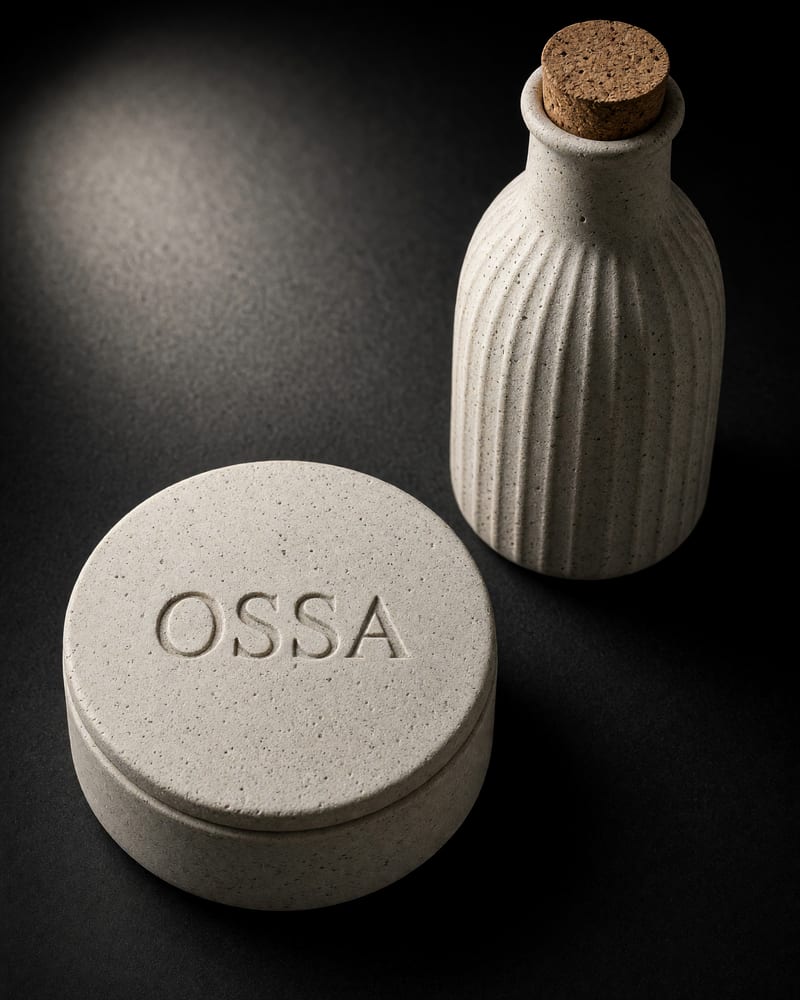 Bone-white unglazed porcelain pill box debossed OSSA beside a ribbed bottle with a cork stopper, lit by a raking spotlight on charcoal