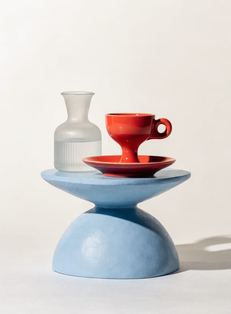Coral espresso cup and frosted glass carafe on a pale blue sculptural plinth.
