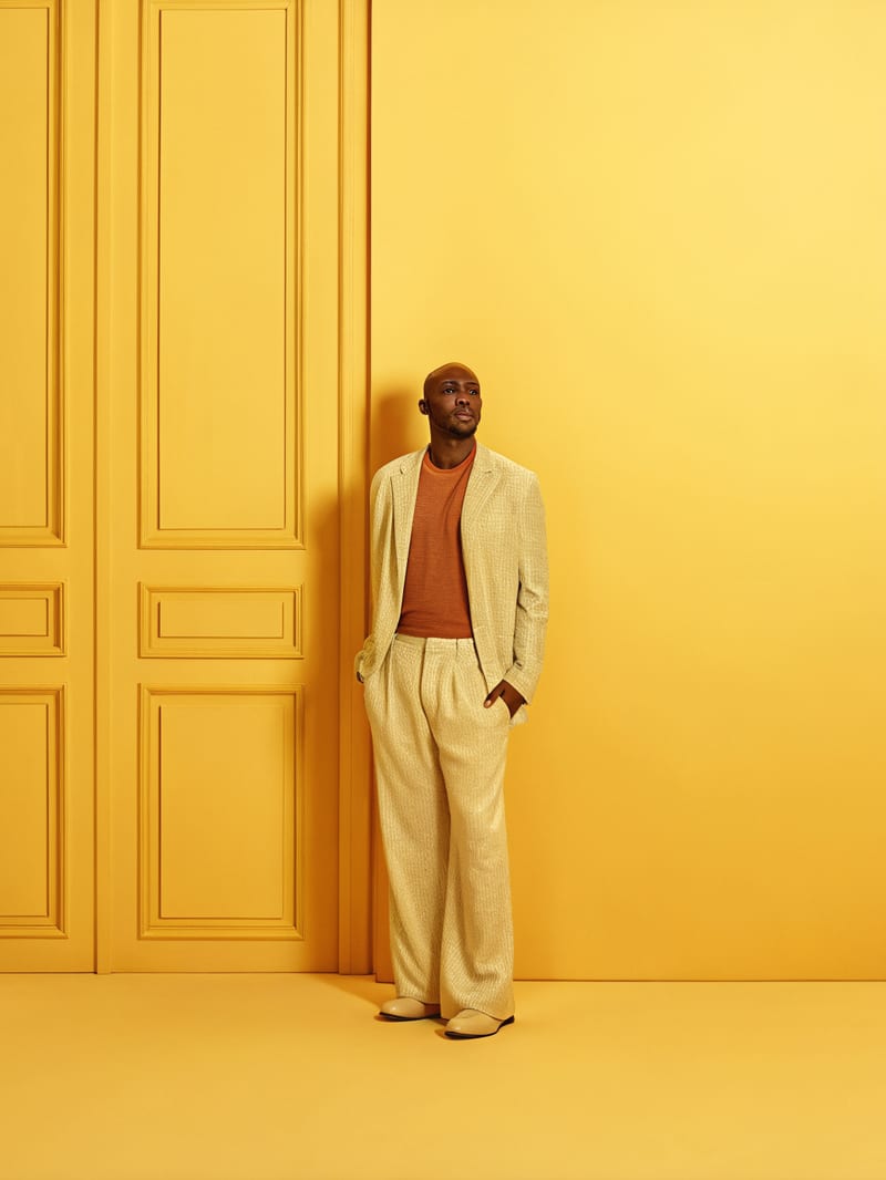 Fashion lookbook image of a man in an ivory suit against a yellow architectural doorway.