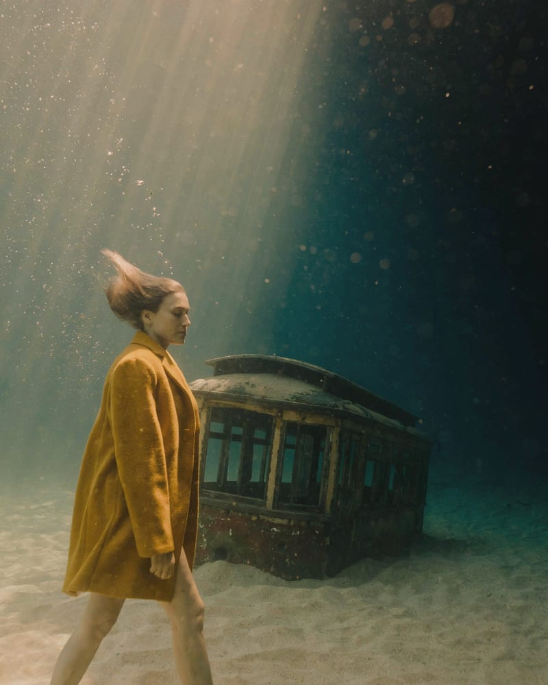 Woman in a mustard wool coat walking on the seabed past a sunken tram, lit by diagonal shafts of sunlight
